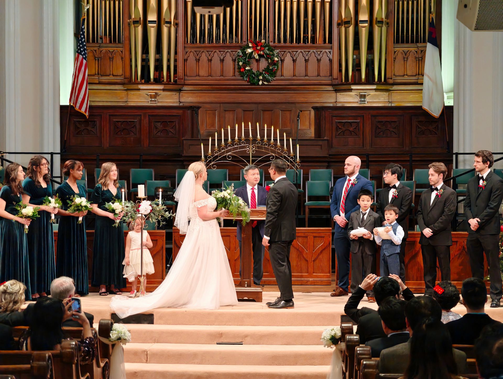 In the Sanctuary in front of the organ, a bride, groom, and party are listening to the pastor preach a wedding sermon.