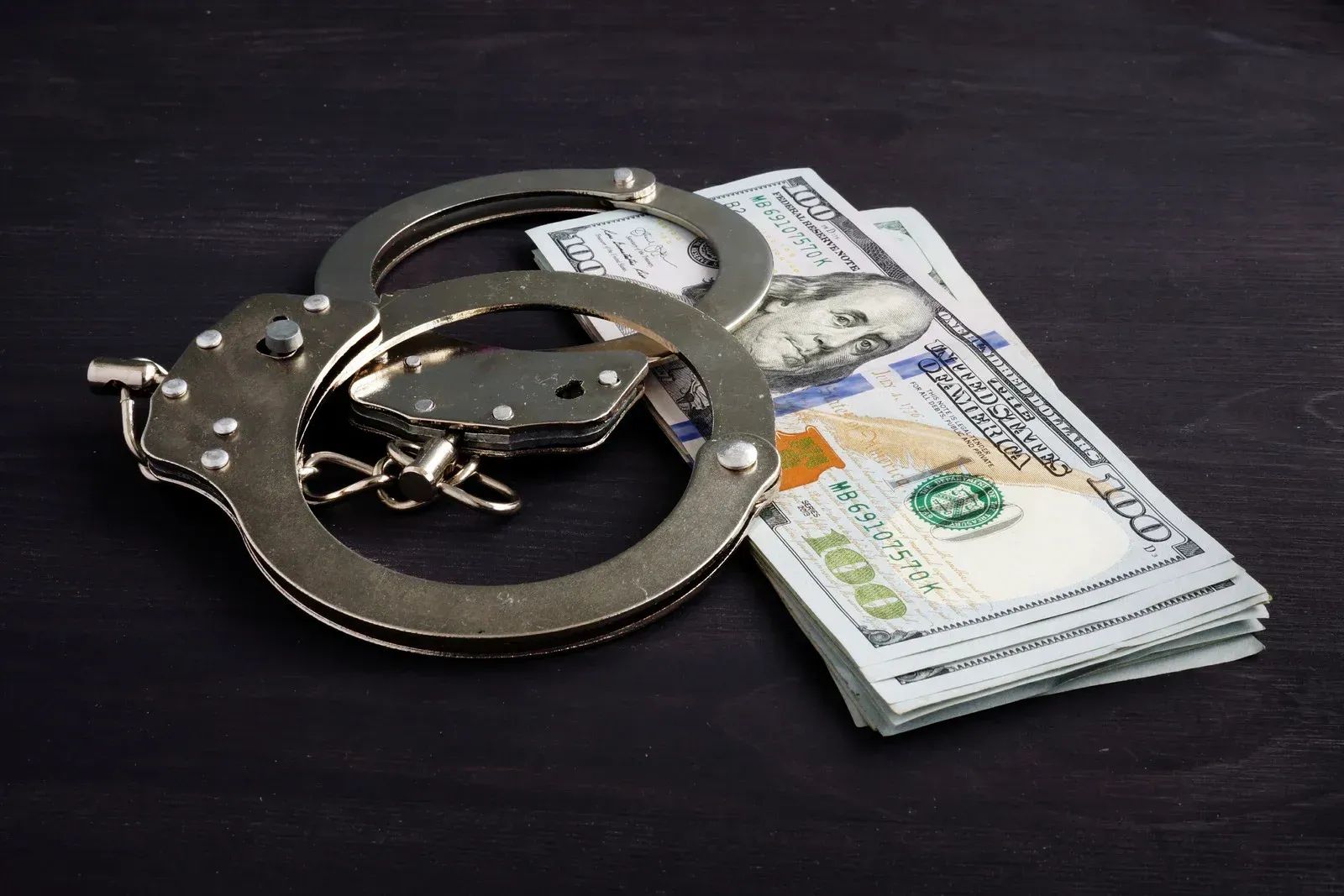 Handcuffs resting on a stack of hundred-dollar bills on a dark surface.