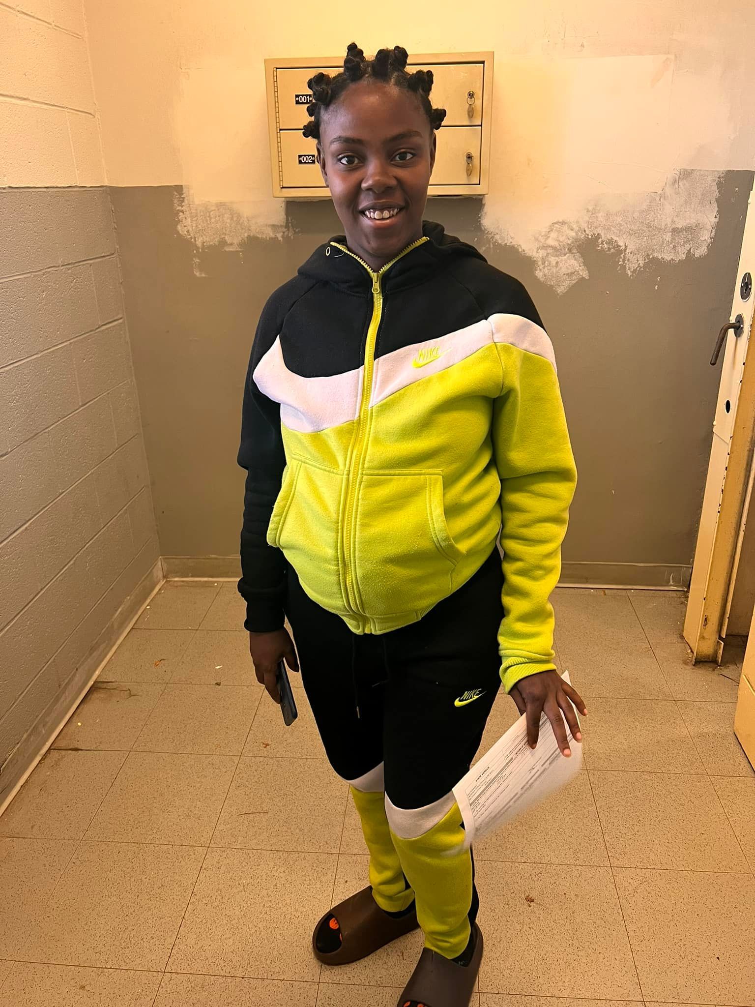 A girl in a yellow and black nike tracksuit is standing in a room holding a piece of paper.