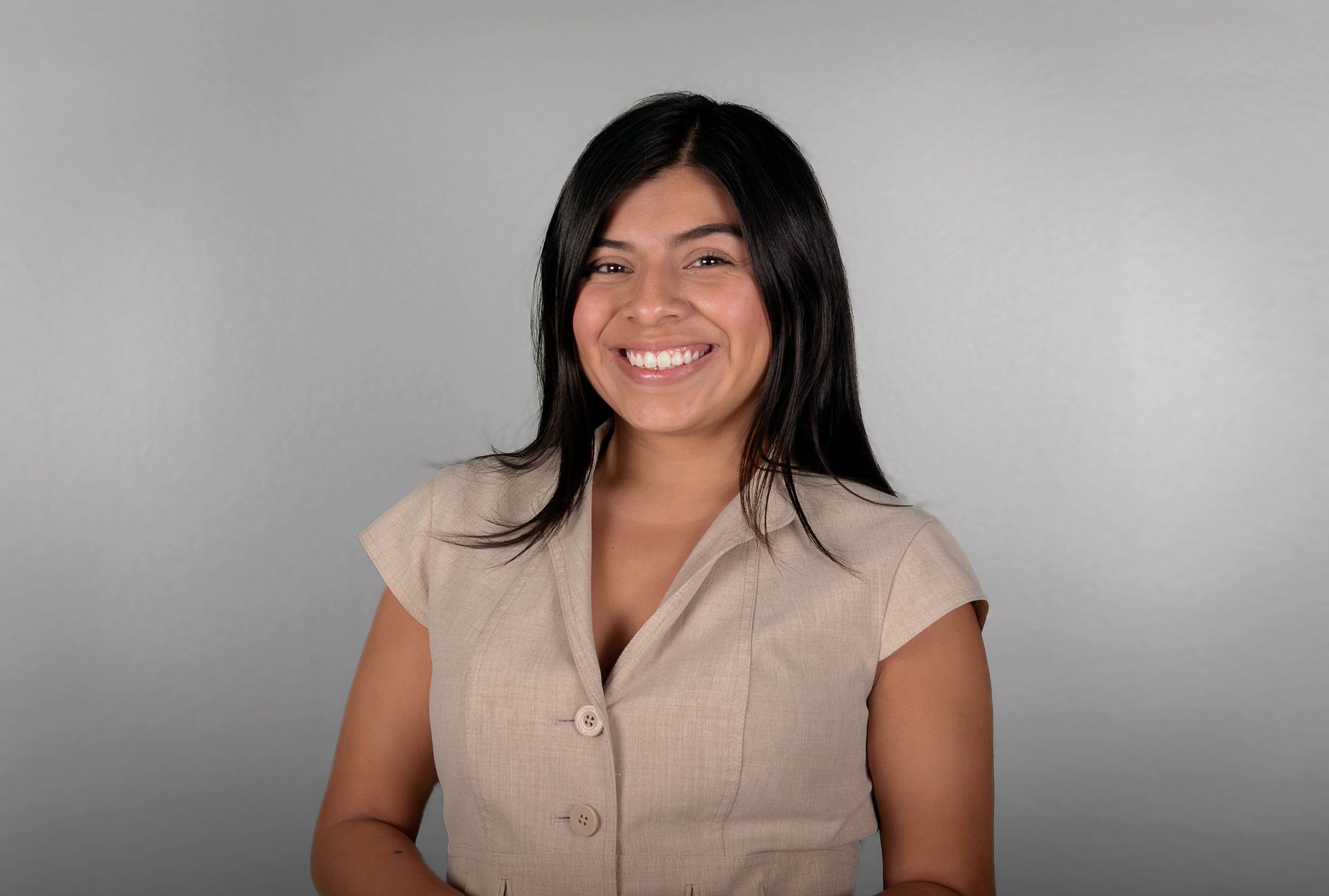 Woman with dark hair smiling in a beige button-up shirt, standing against a grey background.