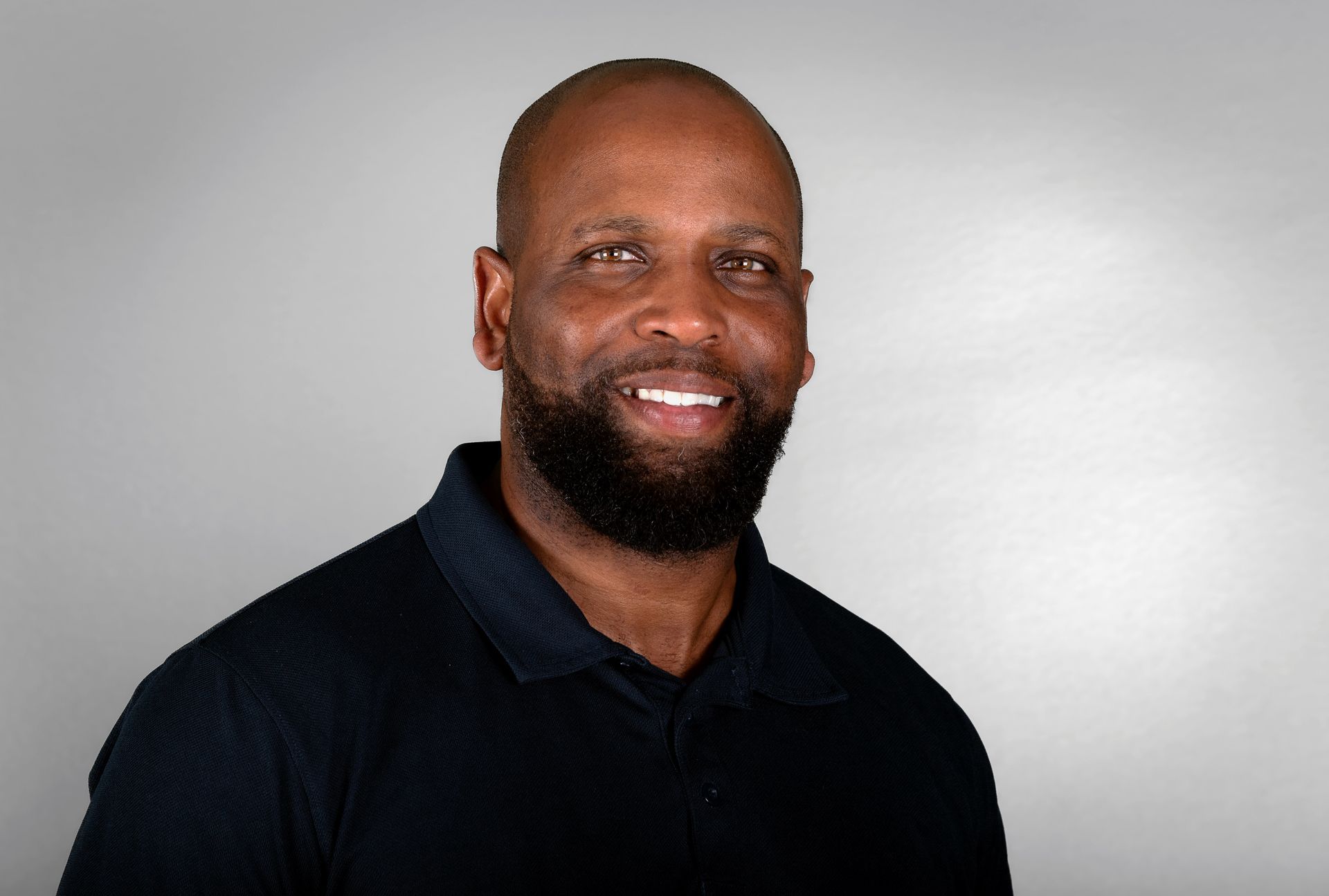 Smiling Black man with a beard, wearing a dark blue polo shirt, against a gray background.