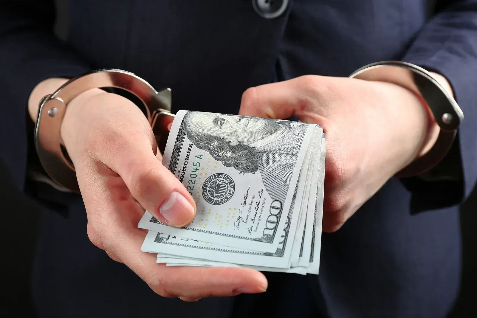 Hands in metal handcuffs holding a stack of one hundred dollar bills against a dark background.