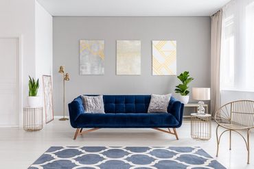 An elegant navy blue sofa in the middle of a bright living room interior with gold metal side tables and three paintings on a gray wall. Real photo