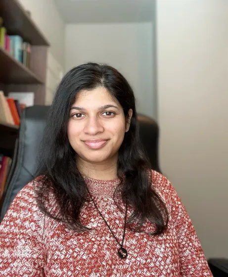 Woman smiling at the camera, wearing a red patterned sweater, dark hair. Bookshelf in the background.