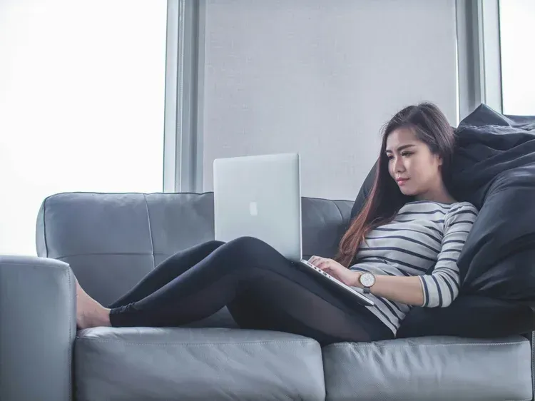 Woman uses a laptop while relaxing on a gray couch in a room with a large window.