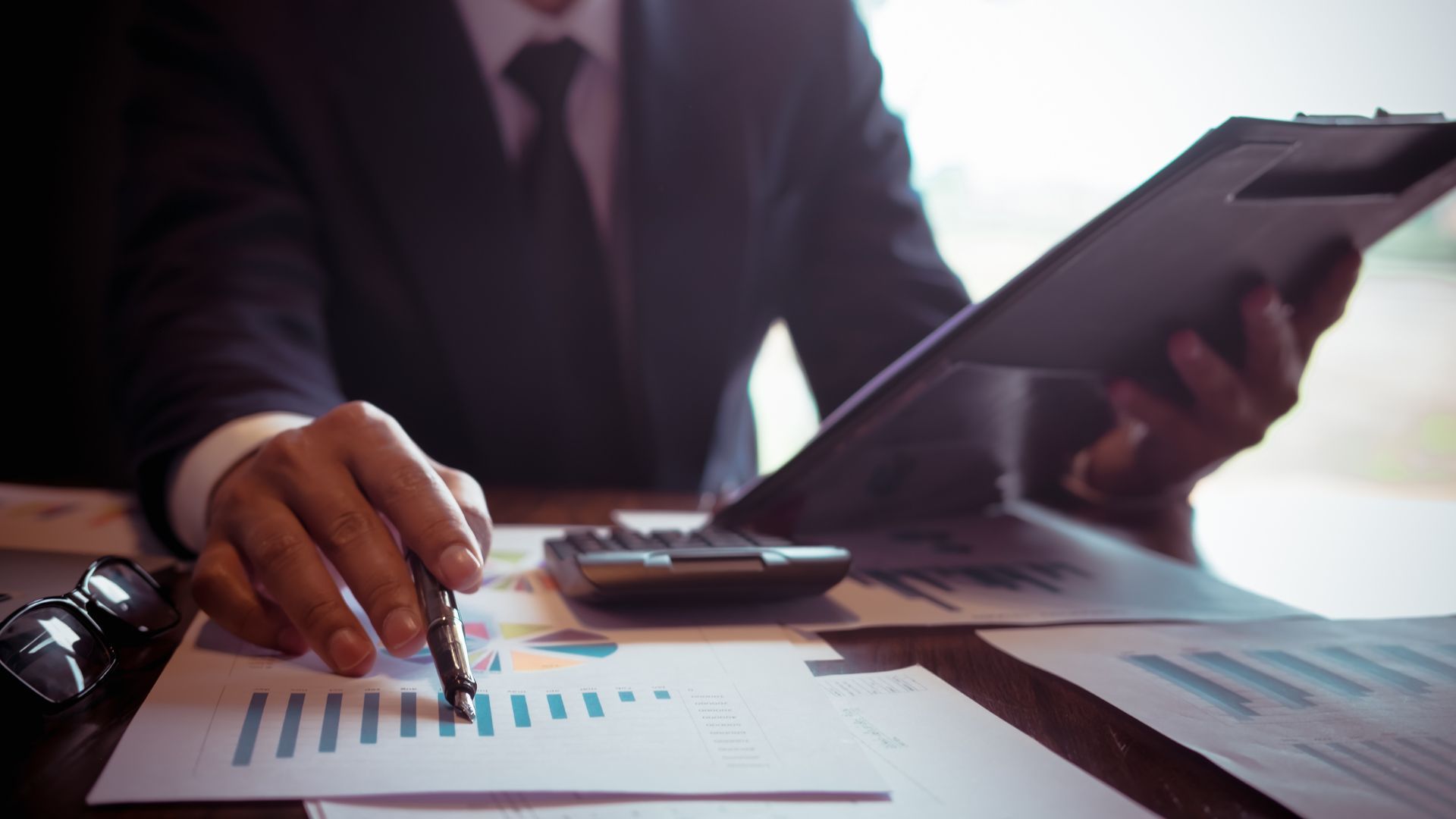 A man in a suit is sitting at a desk using a calculator and a tablet.