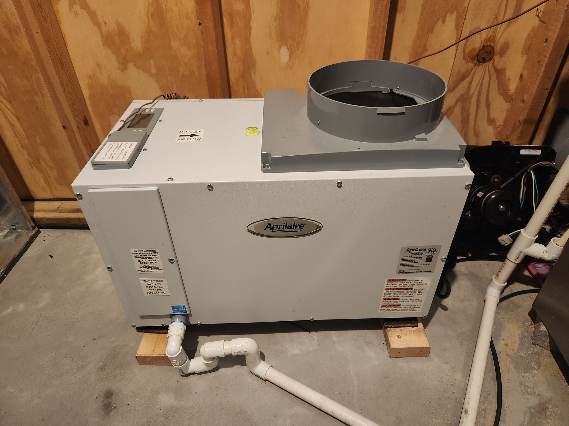 A white HVAC unit in a wooden-paneled room, with ductwork on top and pipes connected at the bottom.