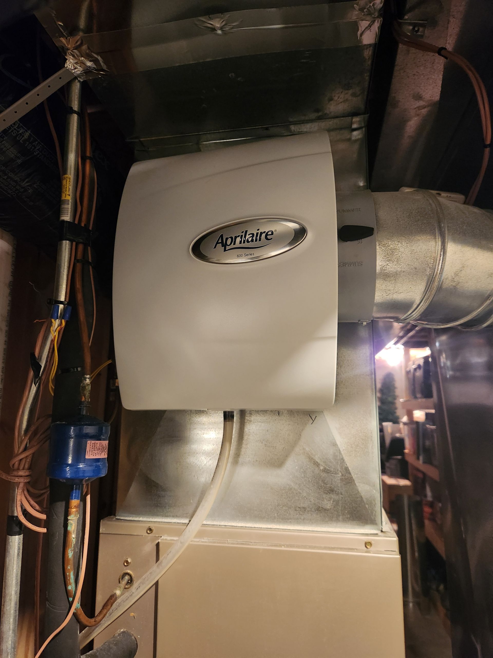 Aprilaire humidifier attached to HVAC ductwork. Beige unit, silver trim, in utility room.