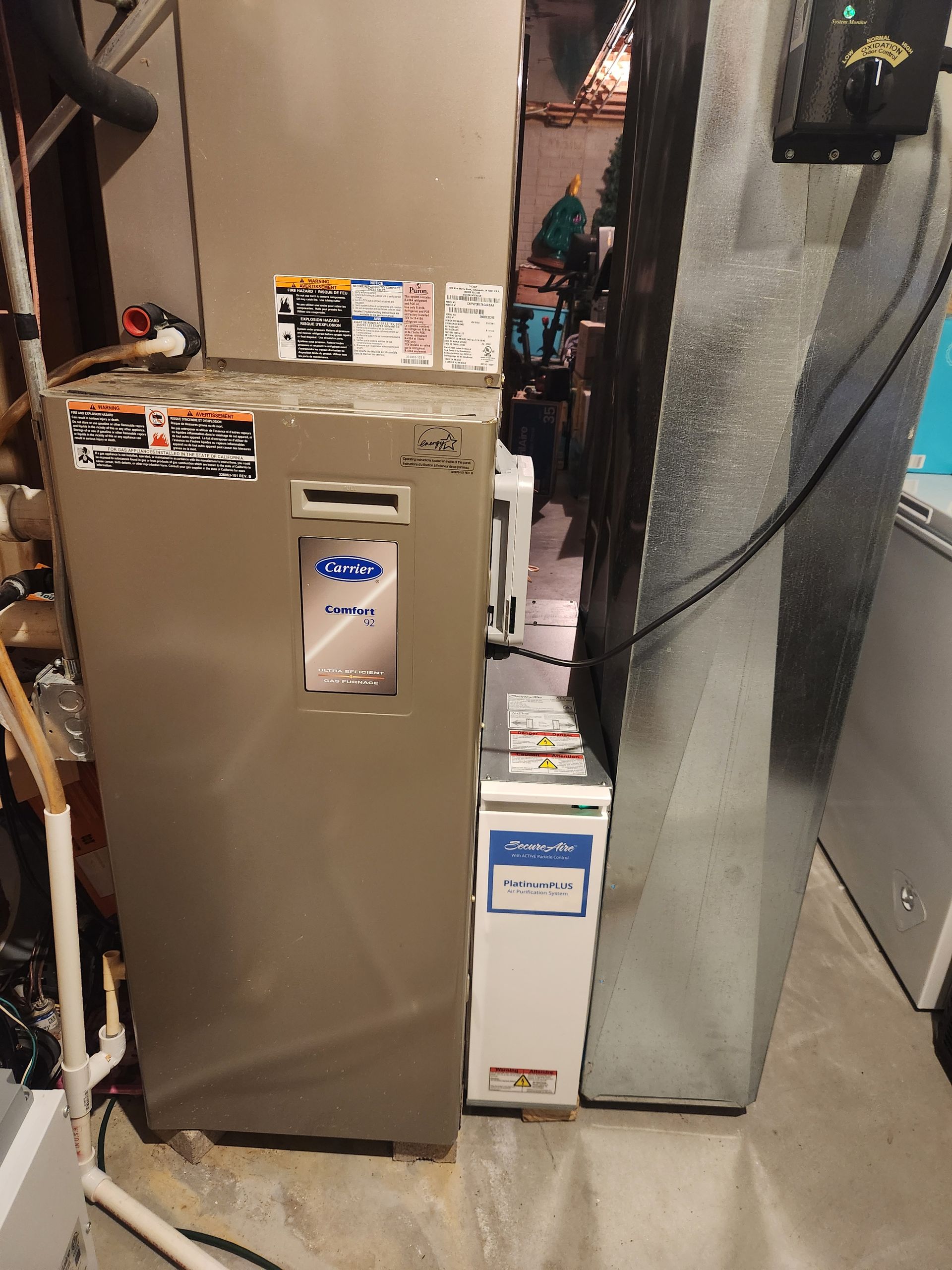 HVAC system in a basement.  A silver furnace is next to a silver air intake.  Piping and a white freezer are also visible.