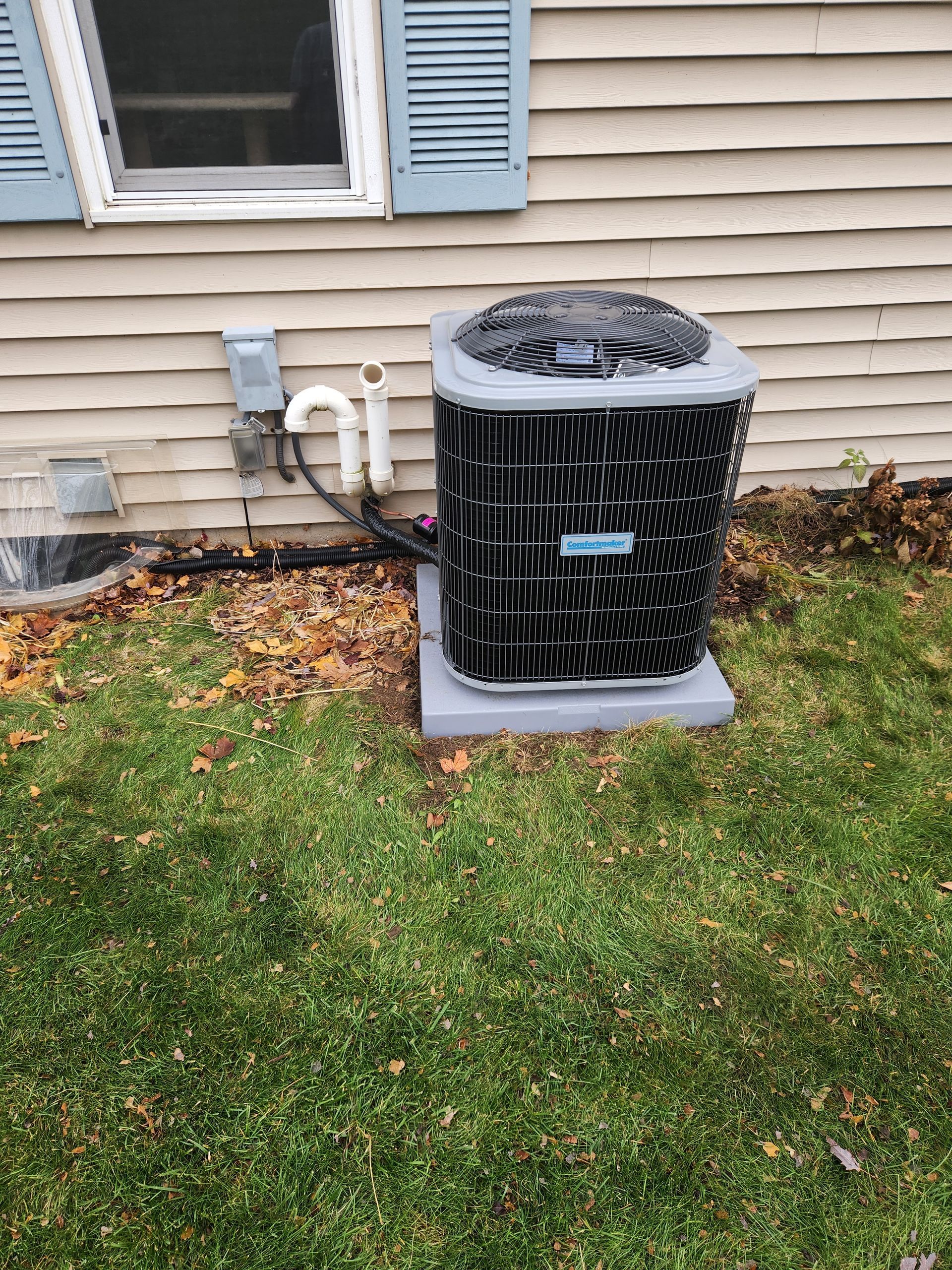 Outdoor air conditioning unit on a concrete pad near a beige house with blue shutters; green grass in the foreground.