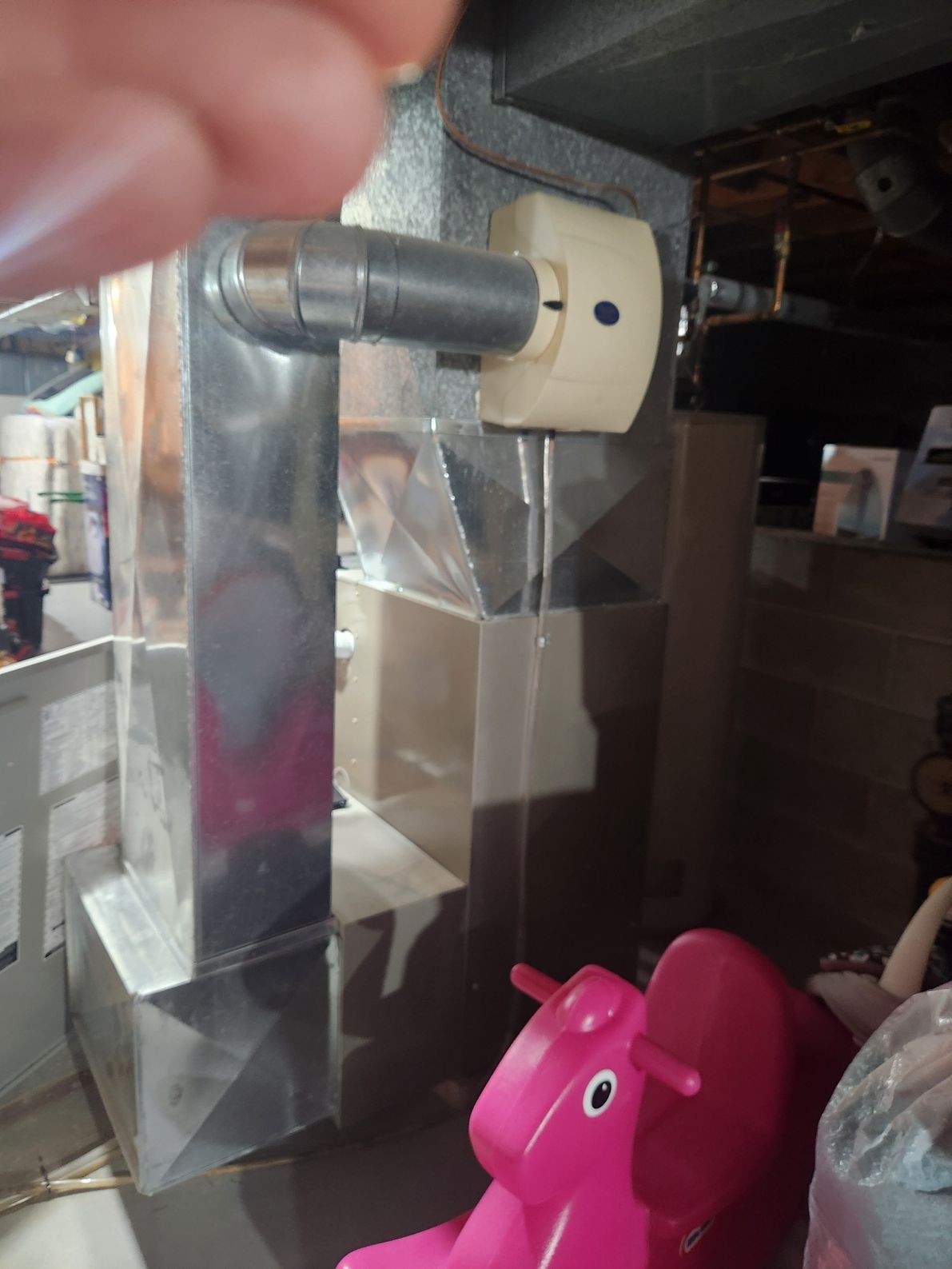 Silver ductwork with an exhaust pipe, cream-colored control box, and a pink toy elephant in a basement.