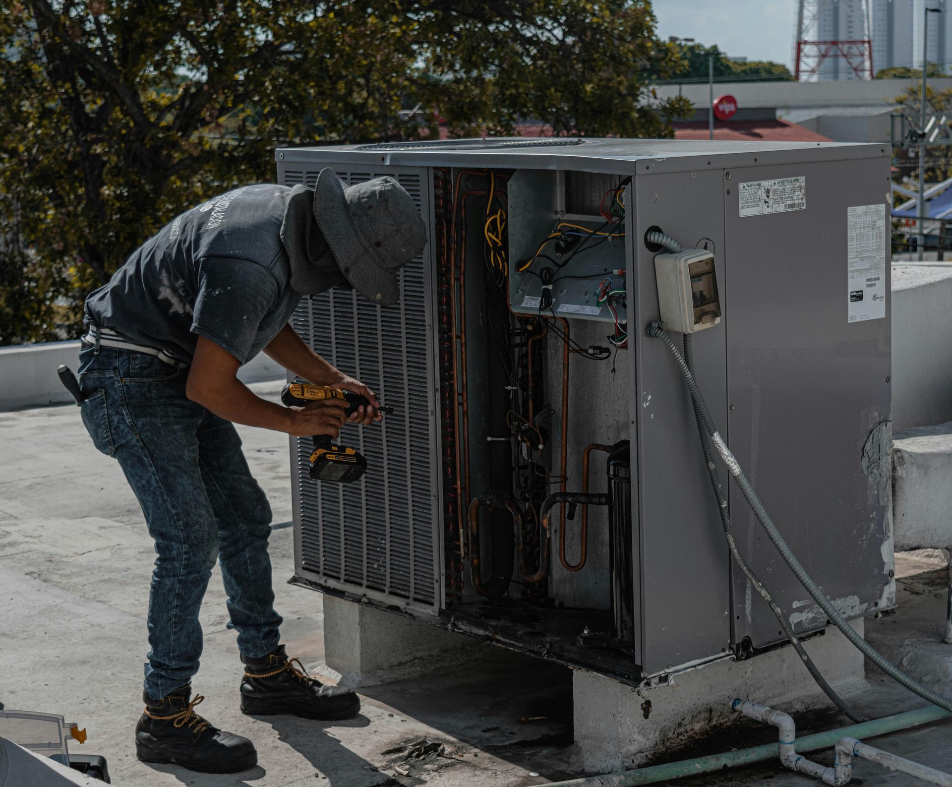 HVAC technician repairs rooftop air conditioning unit with a drill, wearing hat and jeans.