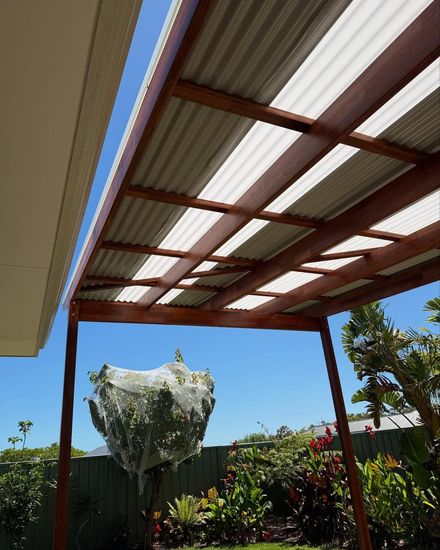 Covered patio with a red metal roof, blue sky, and a hanging white plant basket over a garden