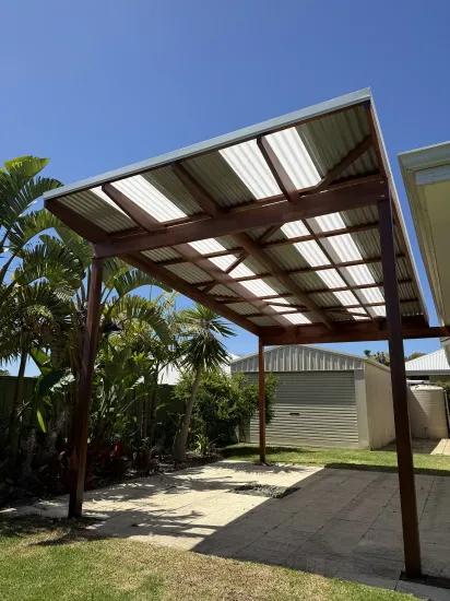 Corrugated metal carport roof over a gravel yard, with trees and a small shed under a blue sky