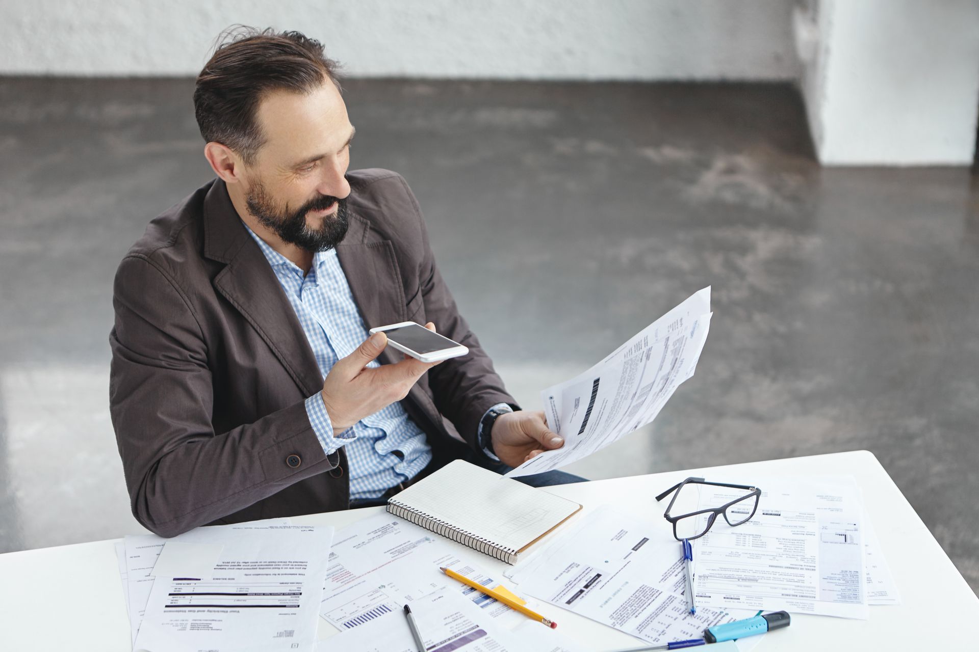 Man in blazer using phone to scan document at a desk with papers, notebook, and glasses.