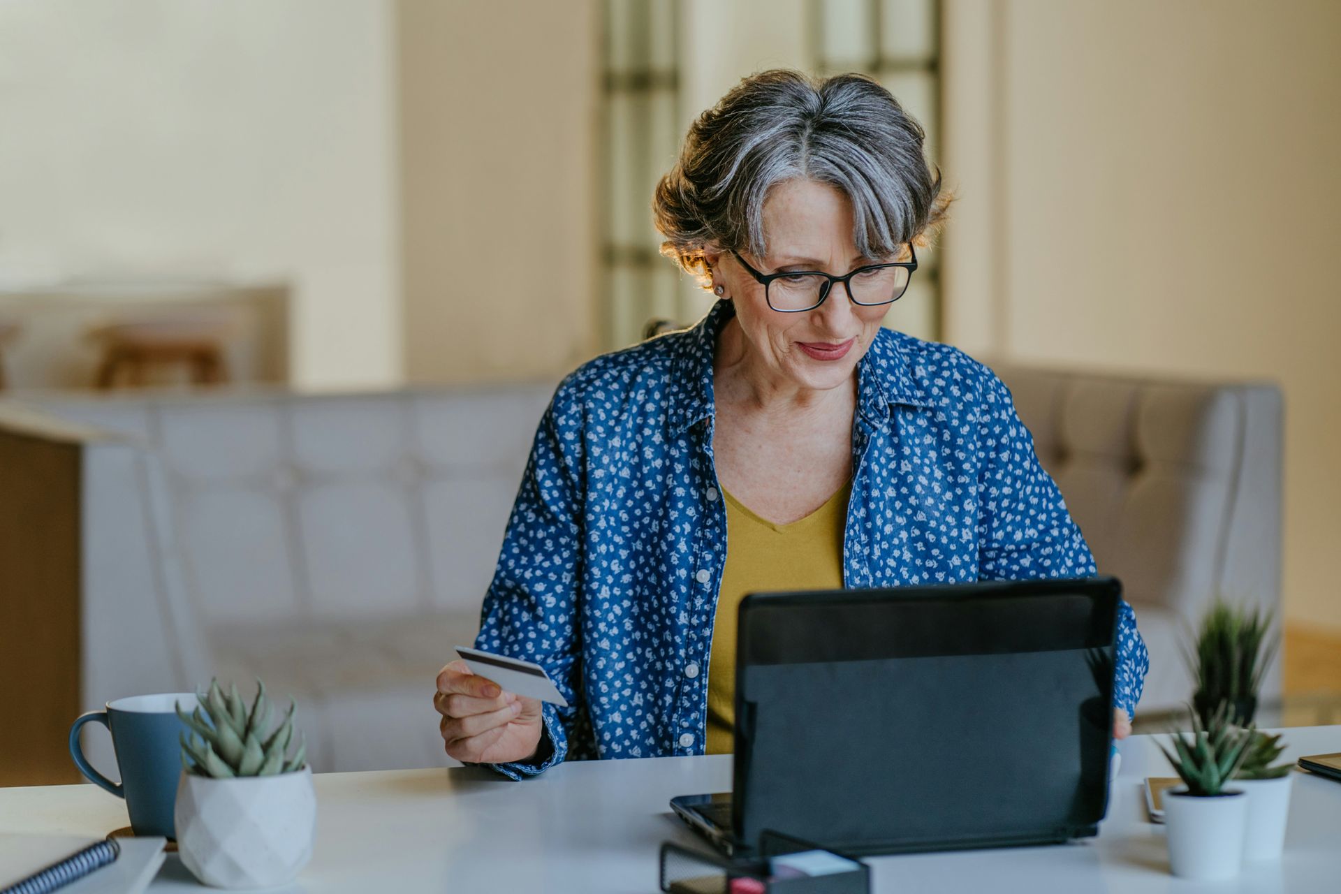 Woman with glasses holding a credit card, looking at a laptop on a desk.