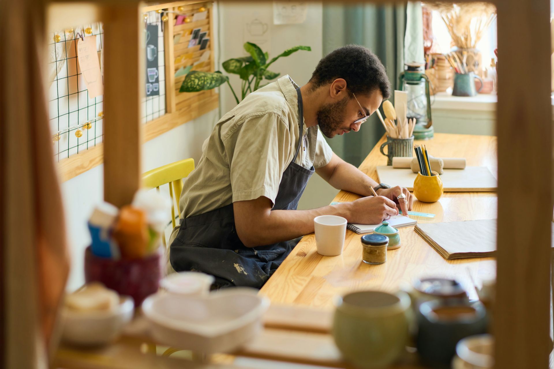 Man in apron, writing at a table with pottery and supplies in a well-lit studio.