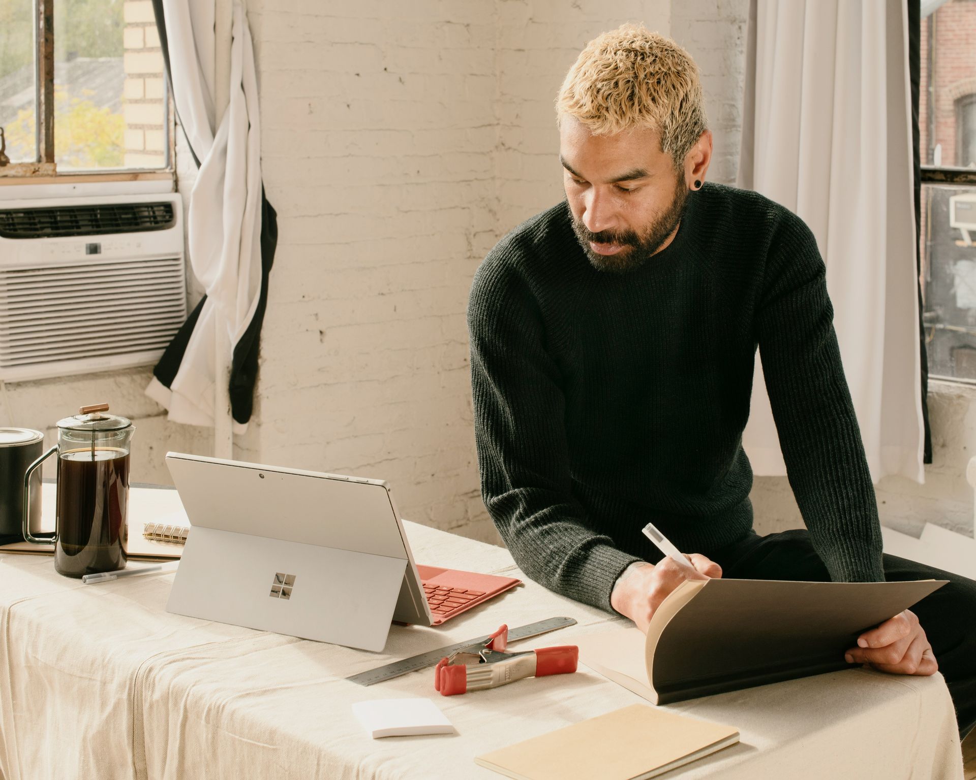 Man sketching at a desk with laptop, coffee, and tools in a bright room.
