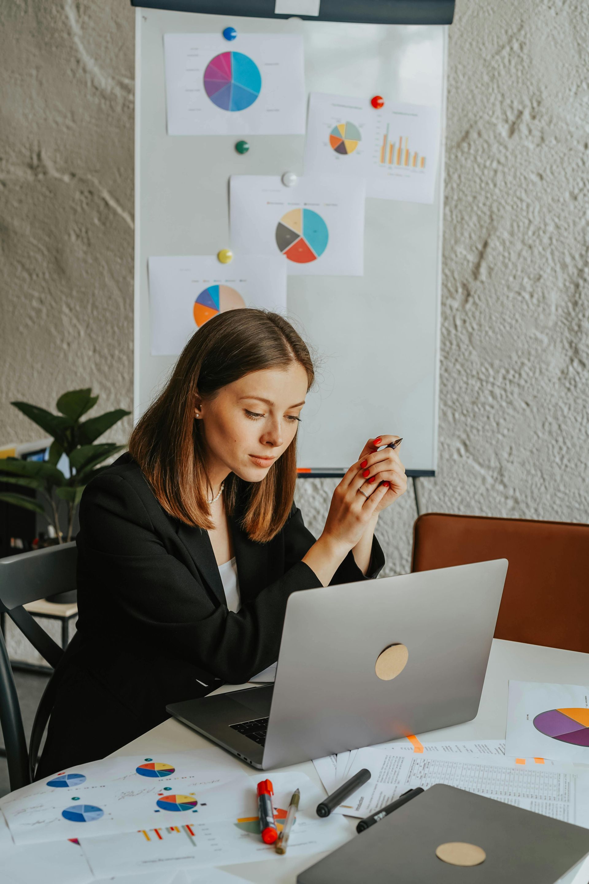 Woman in black blazer working on laptop with charts on whiteboard and table.