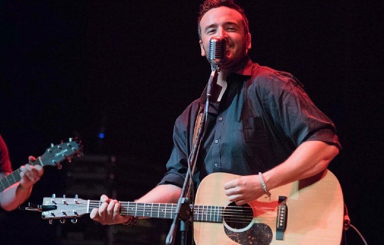 Man on stage singing and playing acoustic guitar; dark shirt, stage lights.