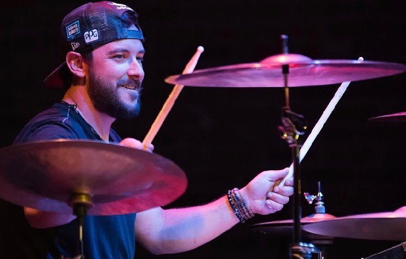 Drummer in cap and t-shirt smiling and playing drums on stage with cymbals and sticks.