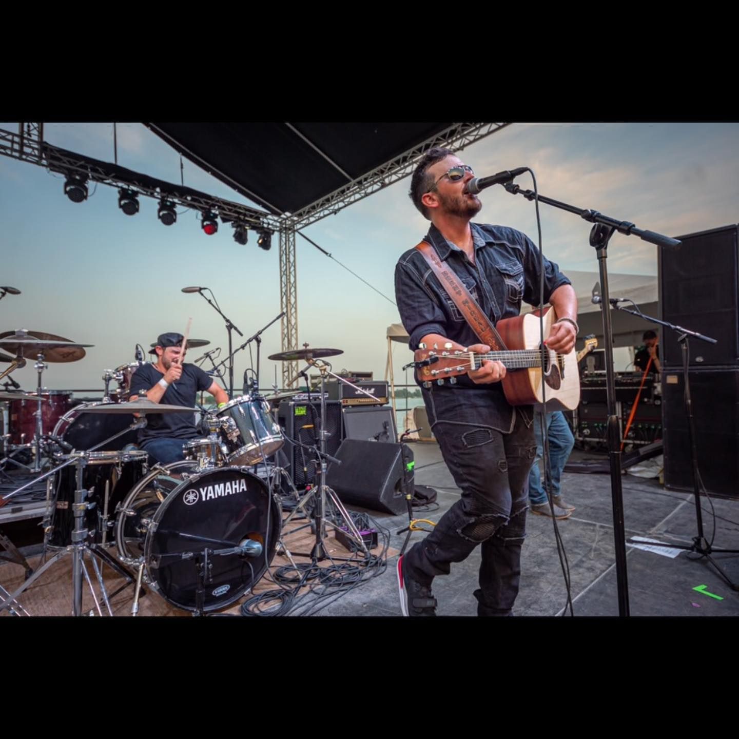 A band performing on an outdoor stage: a singer with a guitar, a drummer, and a guitarist.