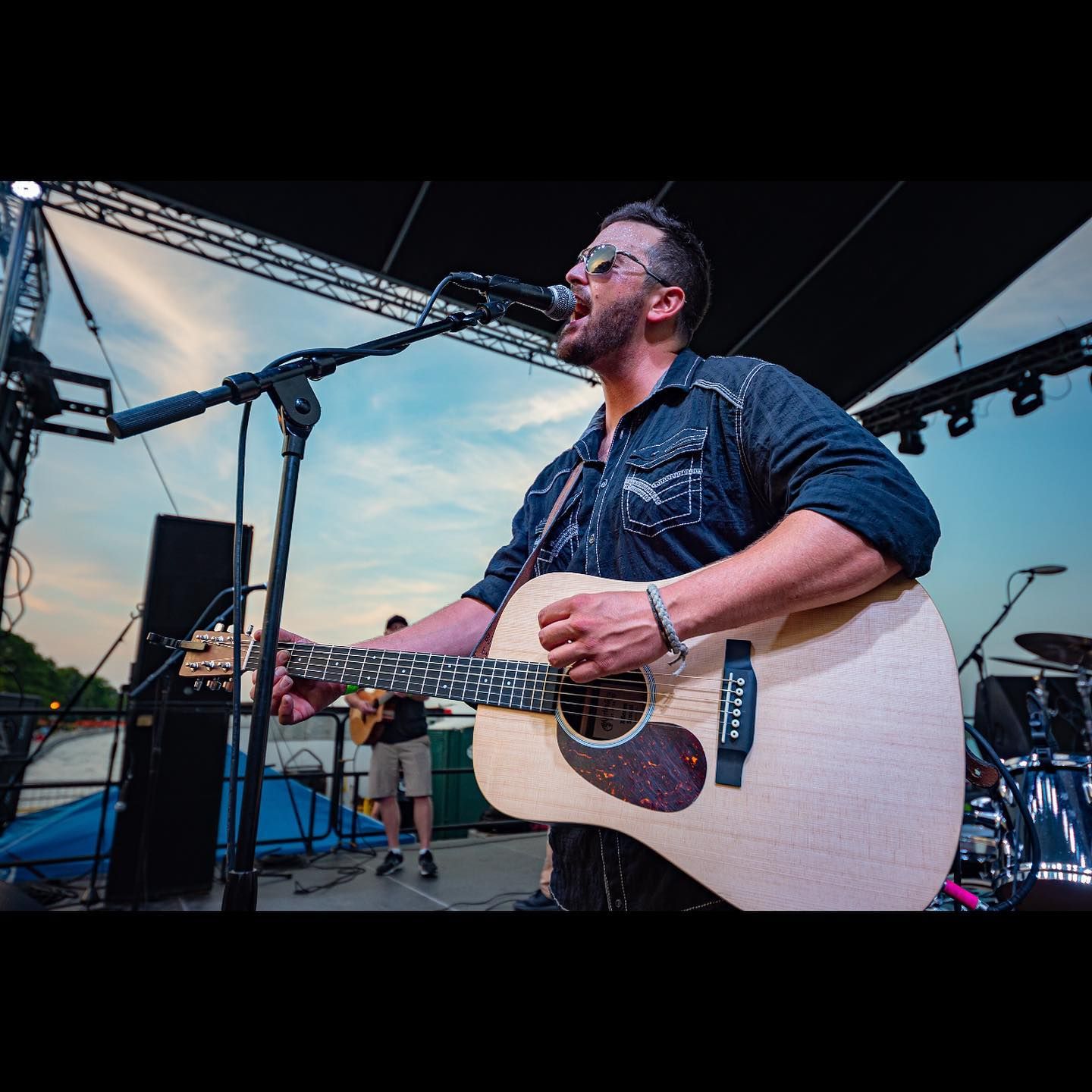 Man playing acoustic guitar and singing at an outdoor concert, wearing a blue shirt, stage and audience visible.