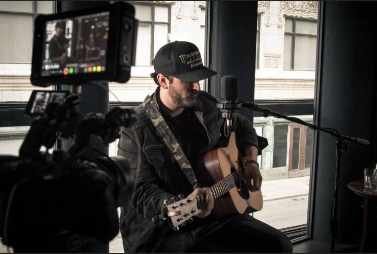 Man playing acoustic guitar in a studio, wearing a cap and jacket, with recording equipment in view.