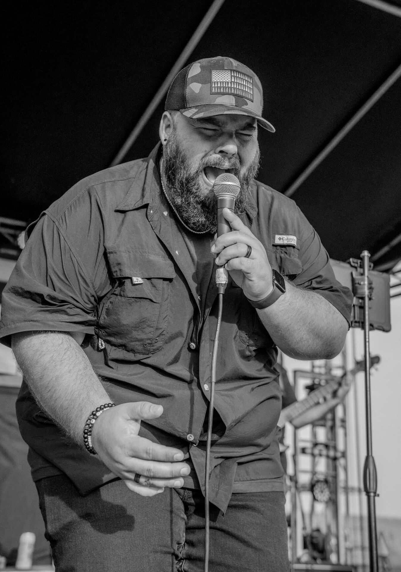 Singer performing on an outdoor stage, holding a microphone and smiling under a canopy.