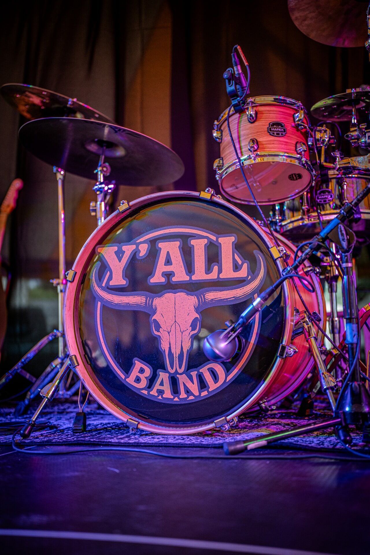 Drum kit with “Y’all Band” logo on the bass drum, lit in purple and pink on stage.