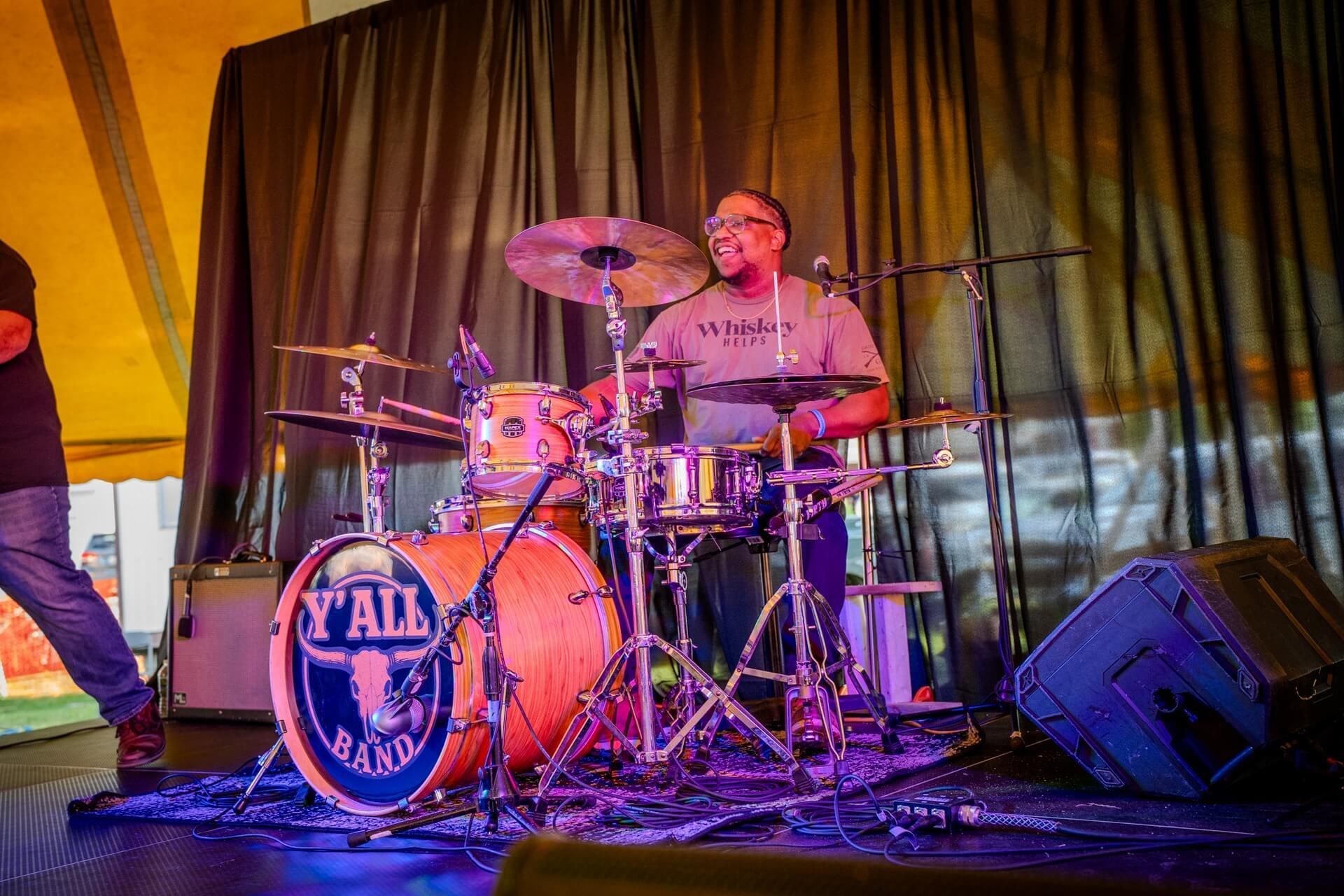 Drummer performing onstage under purple lighting, seated behind a drum kit with a curtain backdrop.