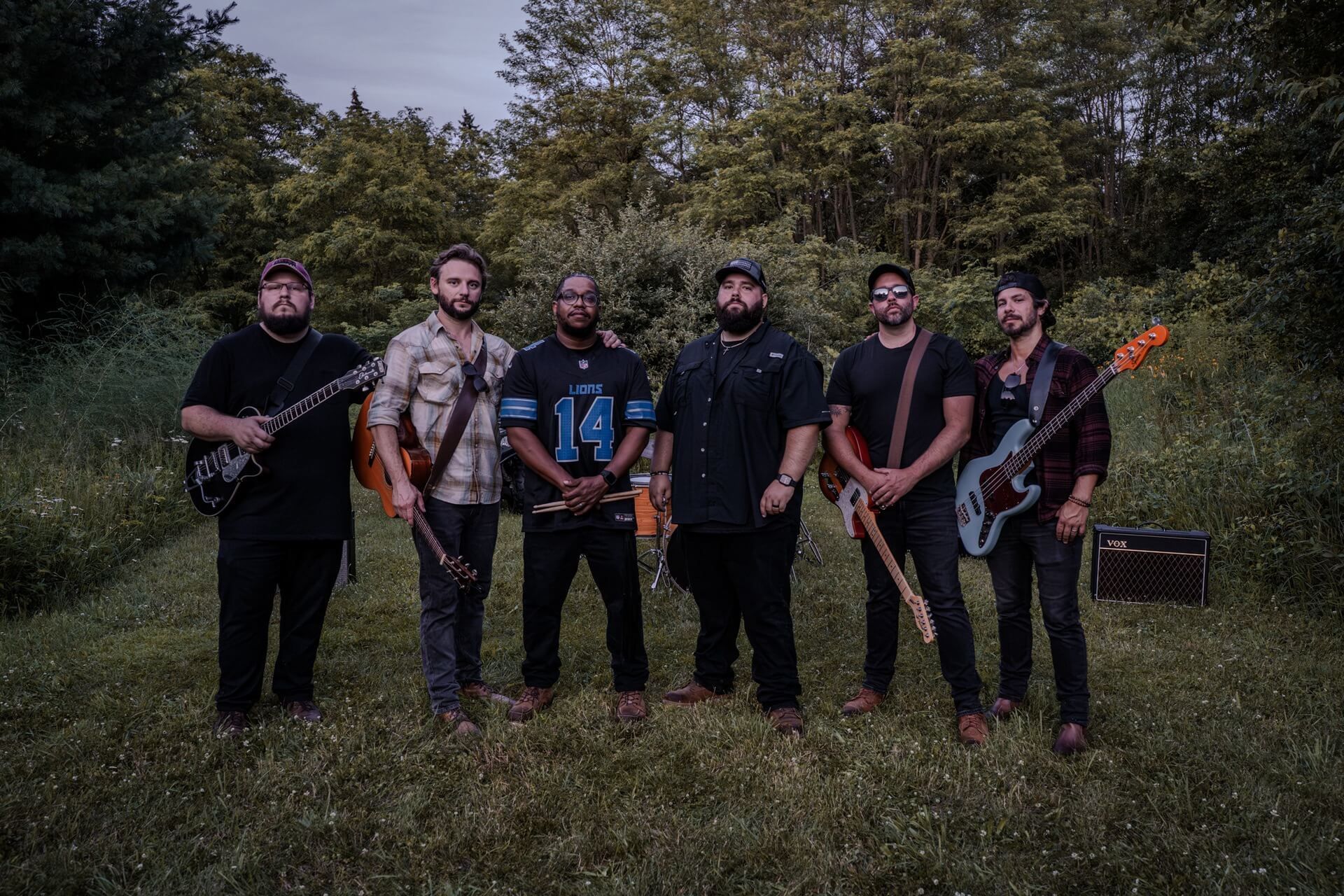 Six musicians standing outdoors in a grassy field with guitars and a bass, under cloudy skies.