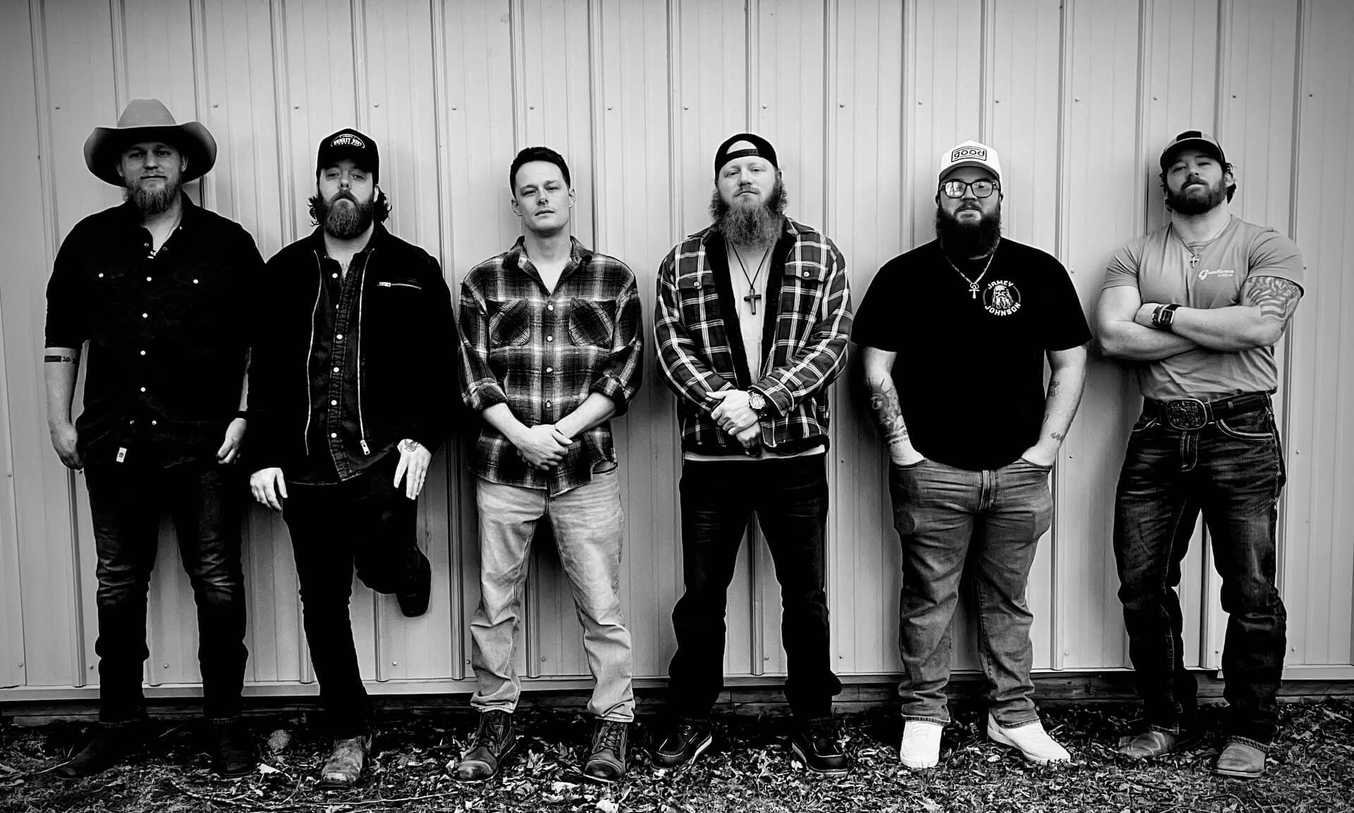 Six men standing against a wooden wall, dressed in casual dark clothing and boots.