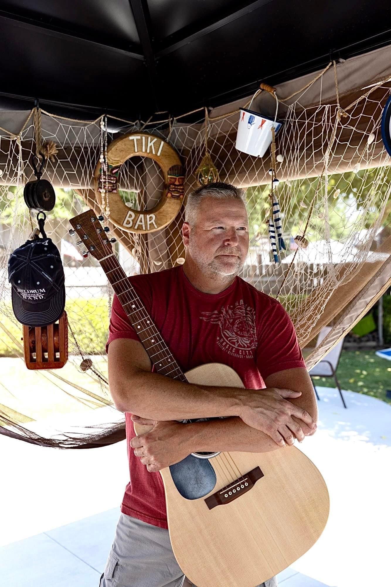 Live band performing on an outdoor stage, with singer-guitarist at mic and drummer behind him.