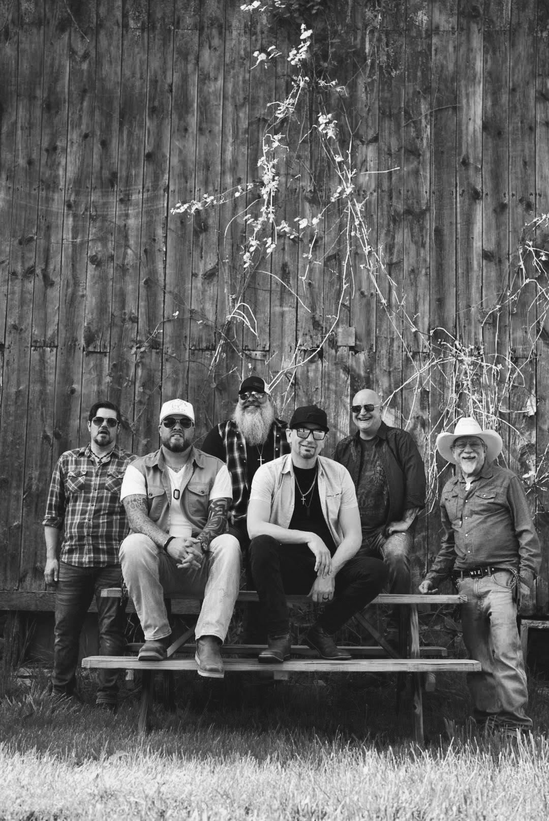Family Tradition Band seated on a wooden bench in front of a weathered metal wall with tall grass.