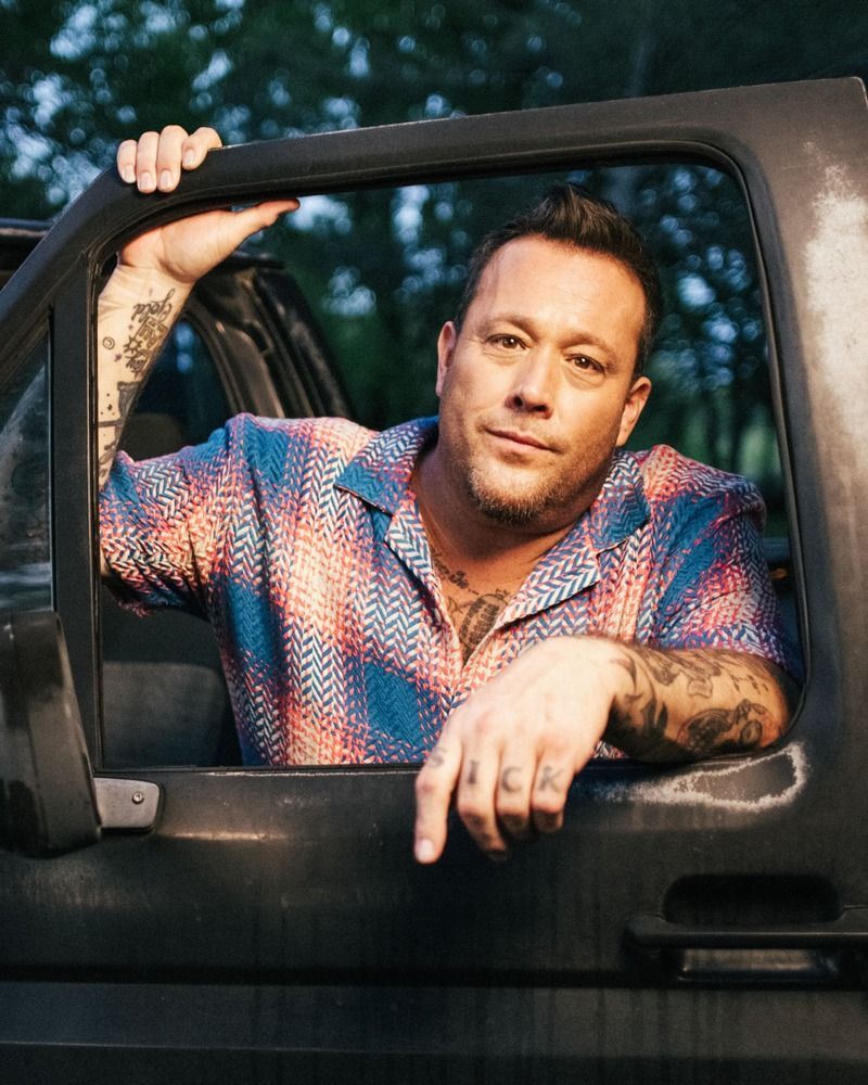 Person leaning on a truck door, wearing a colorful shirt, with tattooed arms and a calm expression.