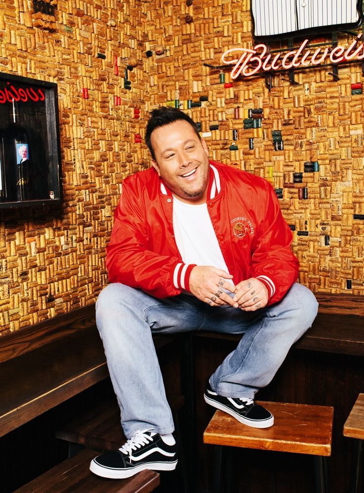 Man in a red jacket sitting on a bench in a dimly lit bar with a Budweiser sign