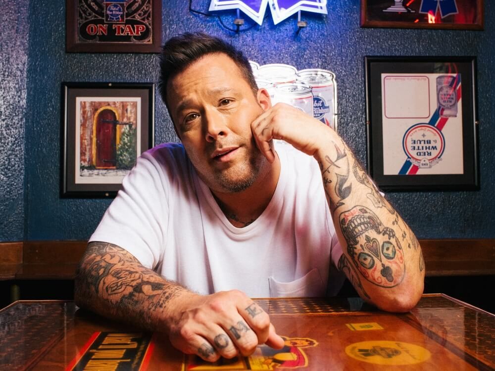 Man leaning on a bar with tattooed arms, wearing a white shirt, in a framed wall display room