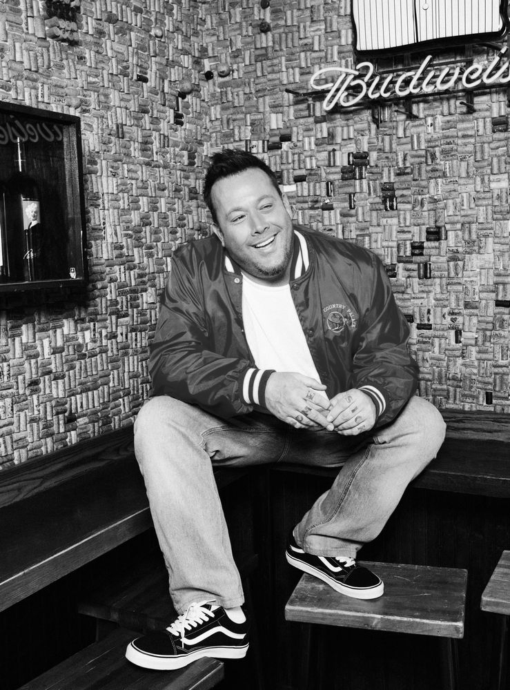 Uncle Kracker is smiling on a bench in a patterned room with a neon sign behind him.