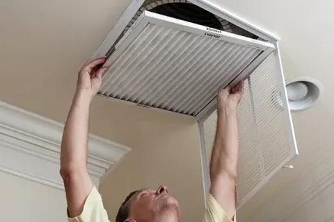 A person in a yellow shirt stands on a ladder, opening a white ceiling vent to replace or inspect an air filter.