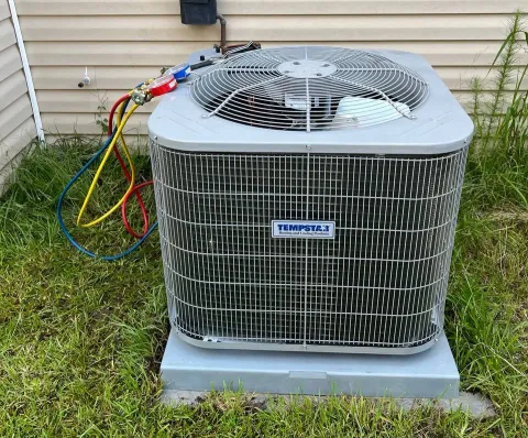 An outdoor air conditioning unit with connected gauges sits on a concrete pad against vinyl siding in a grassy yard.