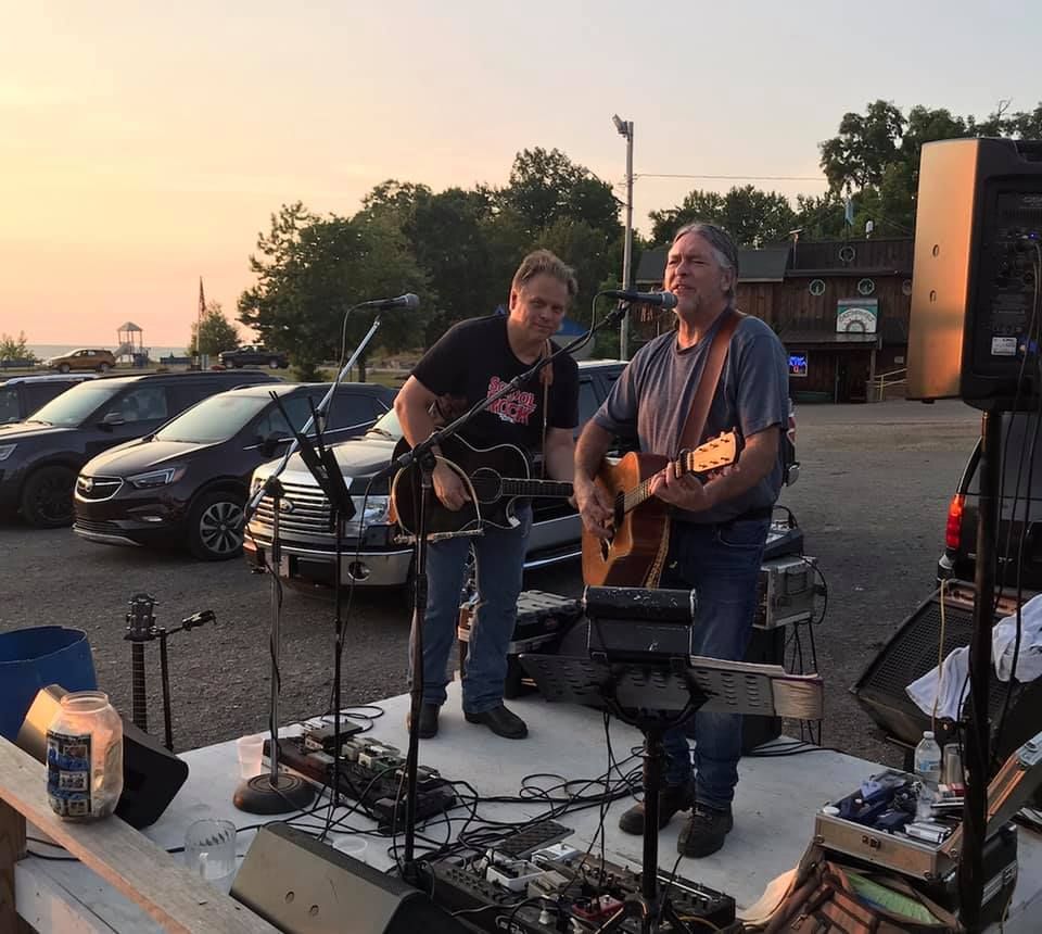 Two men are playing guitars and singing into microphones in a parking lot.