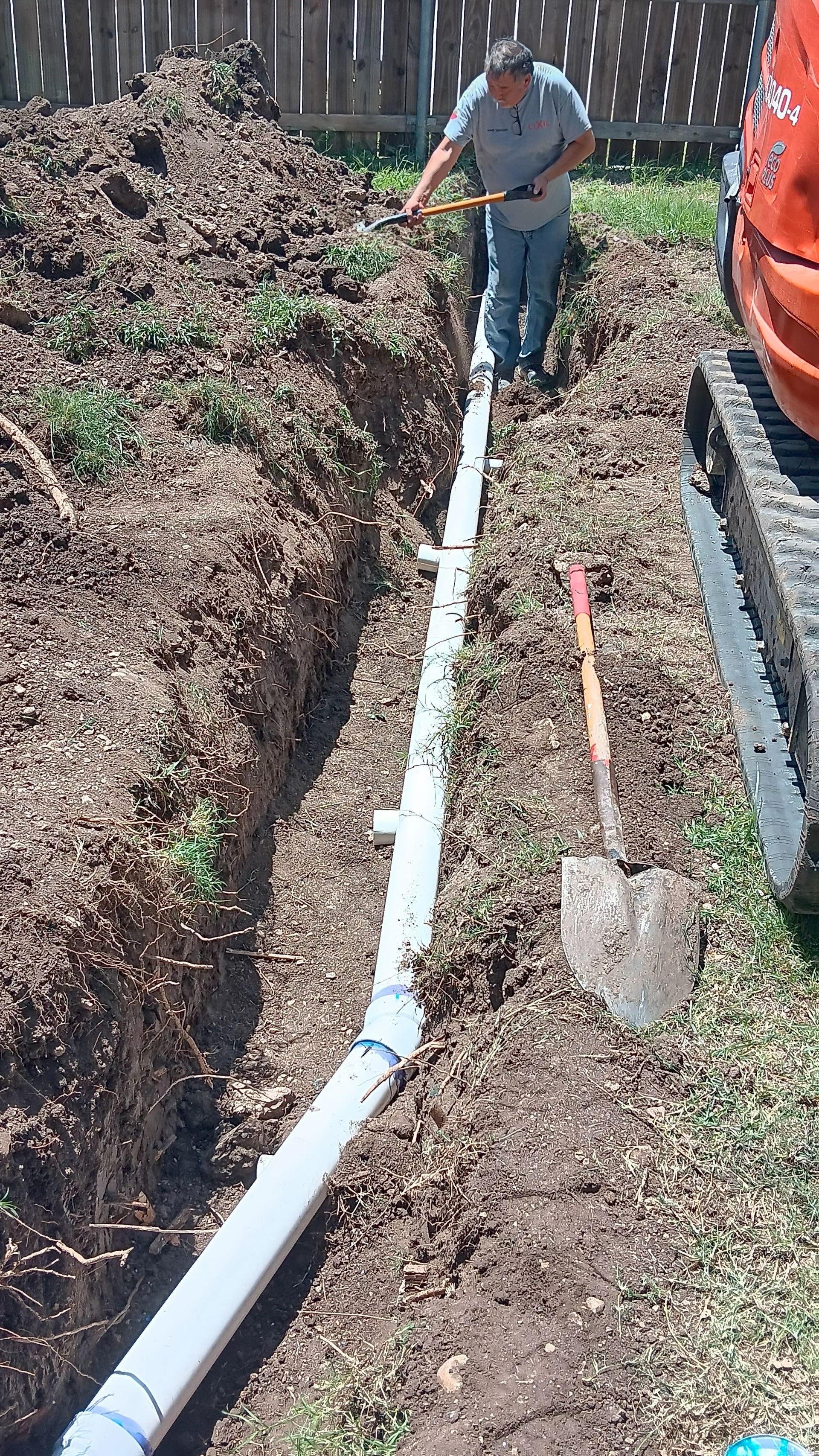 Man installing a white drainage pipe in a trench, outdoors next to a fence and an excavator.