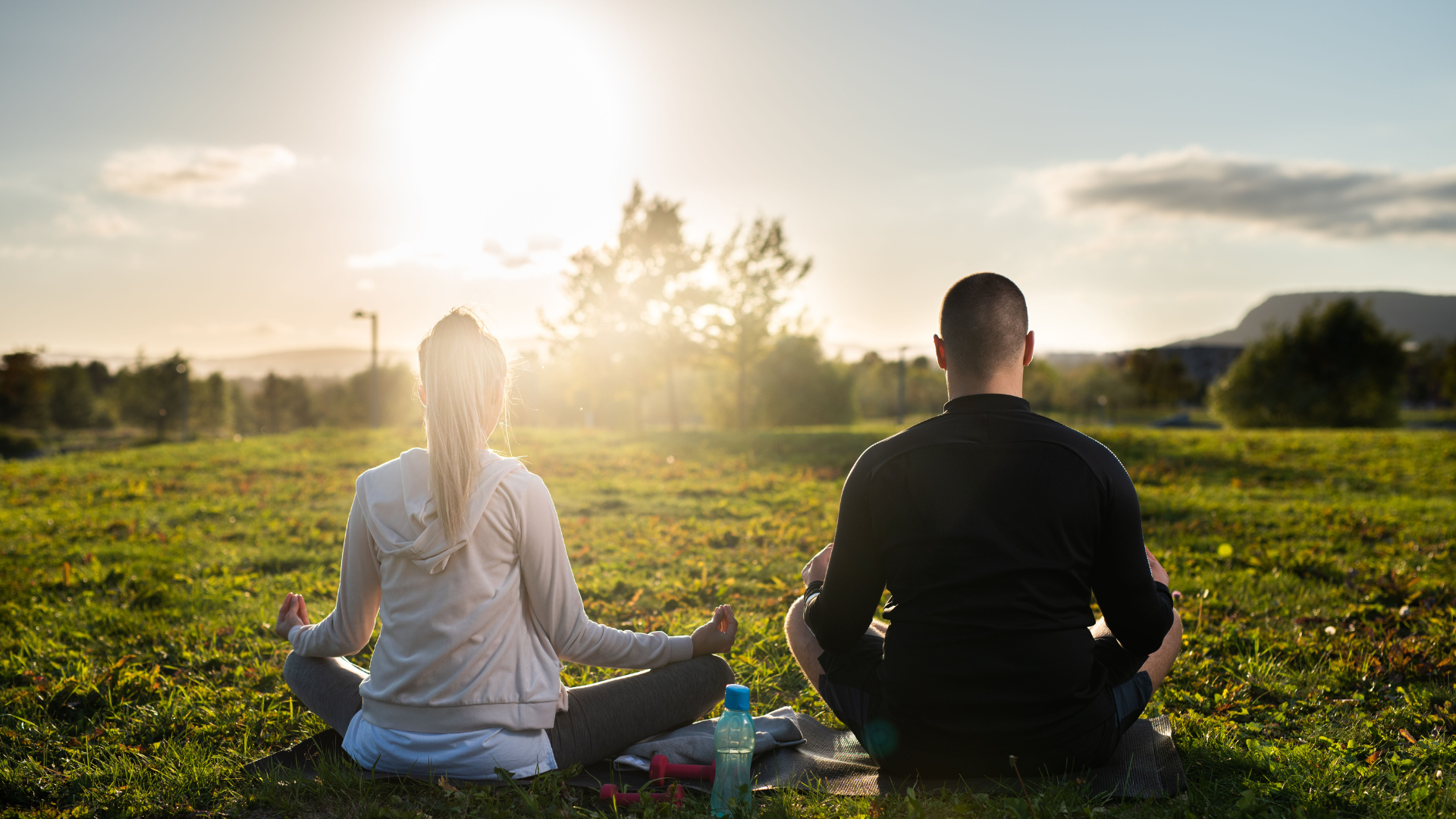 Woman and man in lotus position, meditating in a grassy field at sunset.