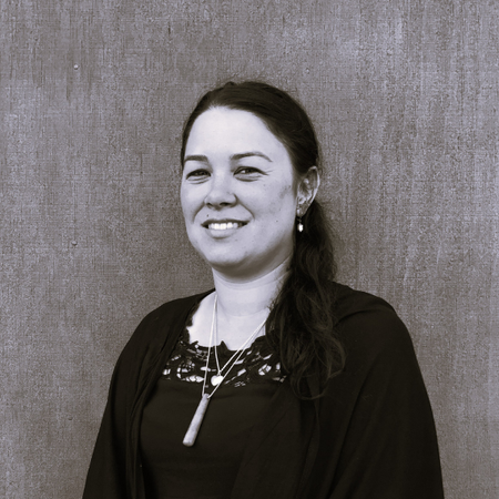 Smiling woman in a dark top with a pendant, standing in front of a wood-grain wall.