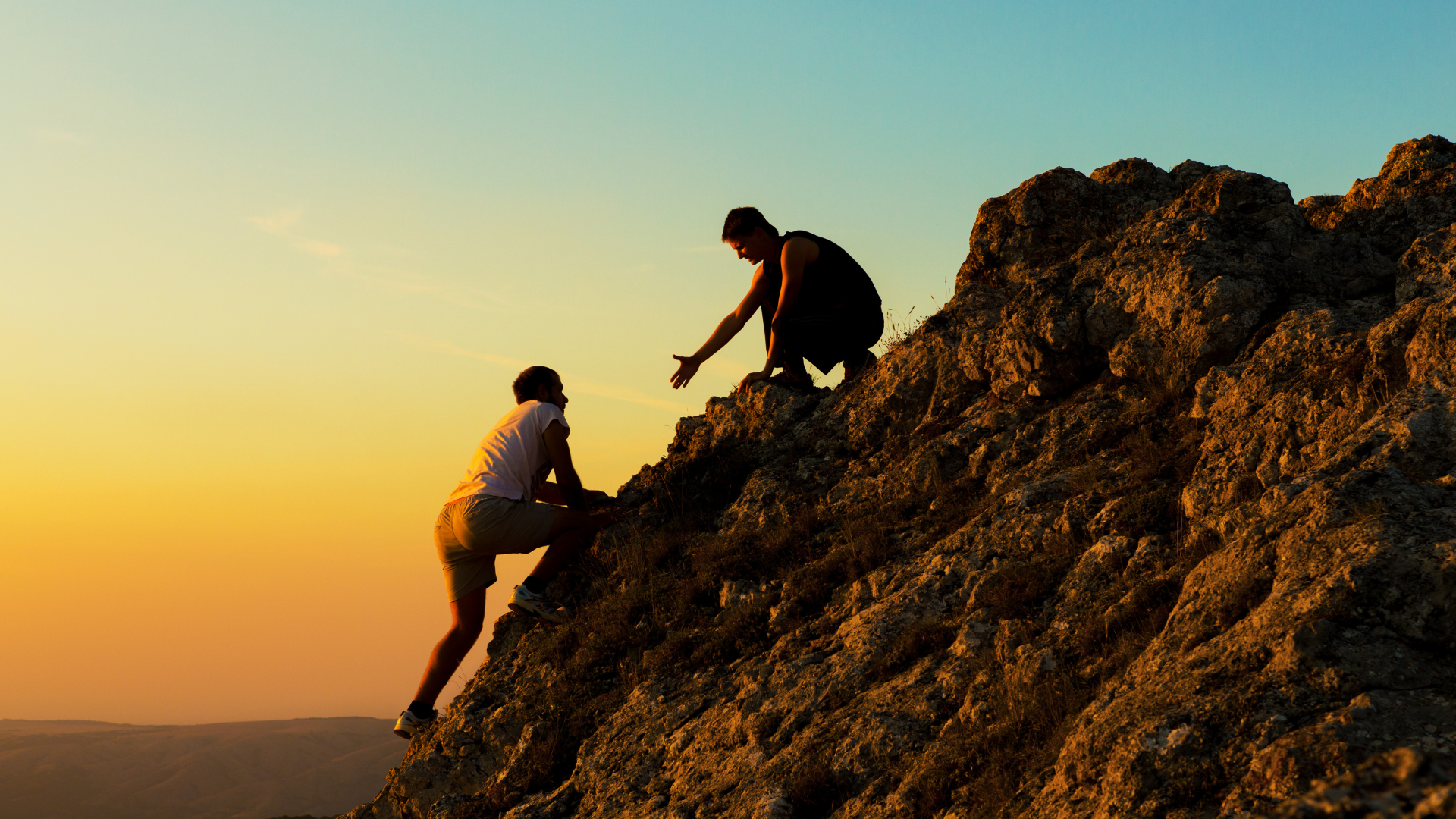 Two people climbing a rocky cliff, one helping the other reach the top at sunset.