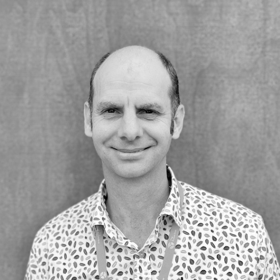 Man with a partially bald head smiling, wearing a patterned shirt and lanyard, in front of a blurred backdrop.