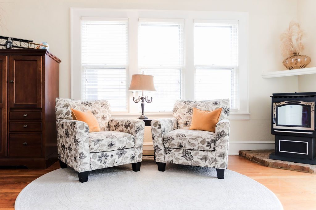 Two floral armchairs with orange pillows, in front of a window, on a white rug.