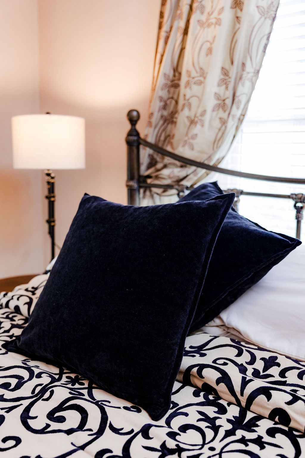 Two navy pillows on a bed with a black and white patterned comforter, lamp, and floral curtains.