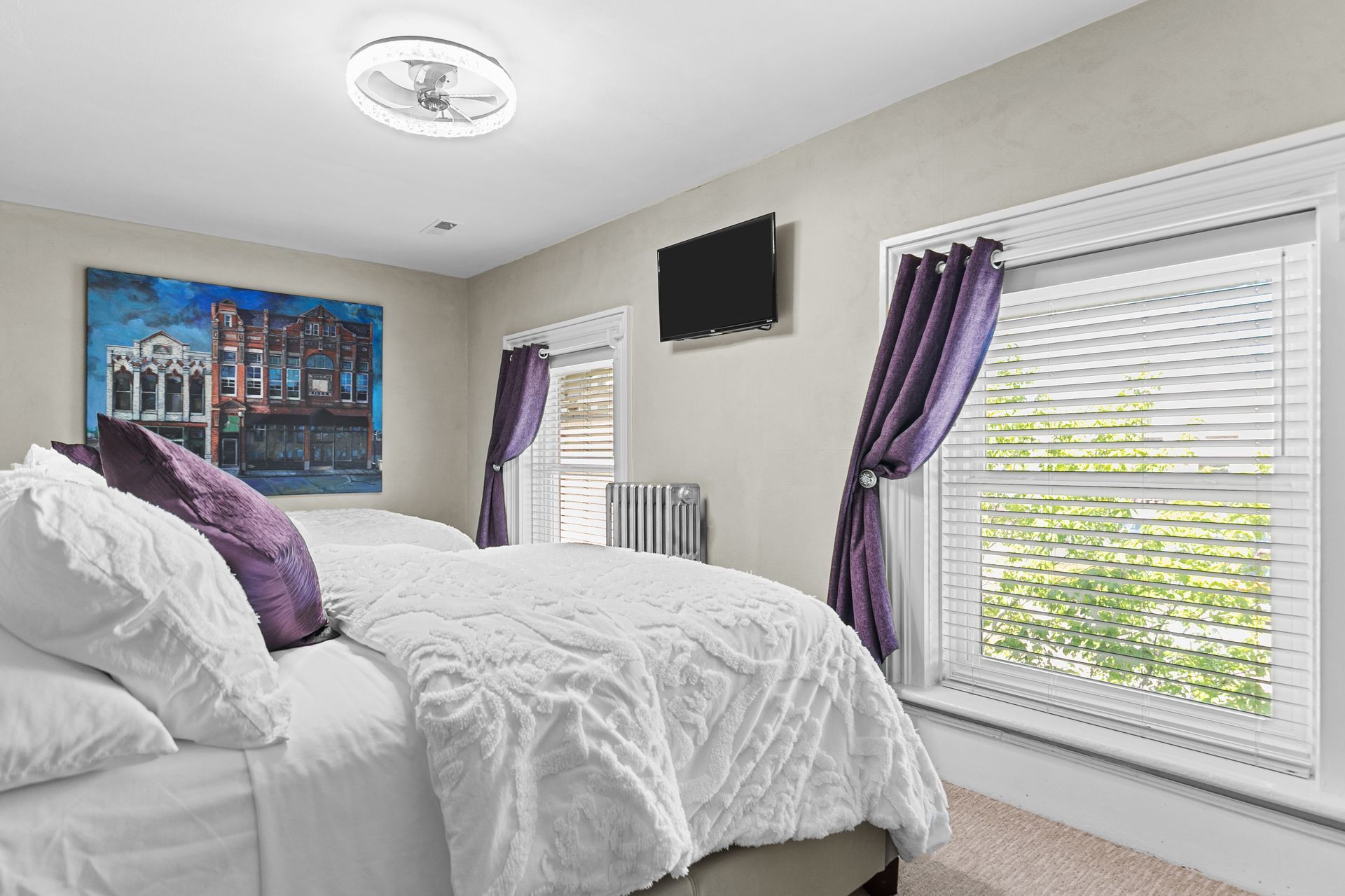 Bedroom with white bedding, purple pillows and curtains, blue and red artwork, and a mounted TV.